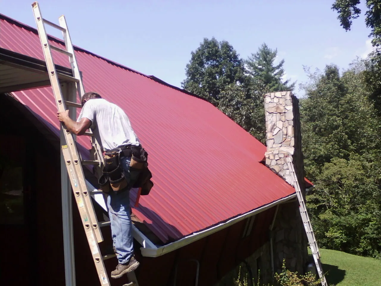 Roofer on a ladder beside a red metal roof and stone chimney — Soffit & Fascia Repair by Roofing Brothers in Brigantine